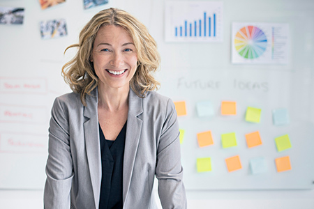 woman-standing-in-front-of-wall-with-charts-and-stickies Woman wearing a suit, smiling in front of a wall with charts and sticky notes on it.